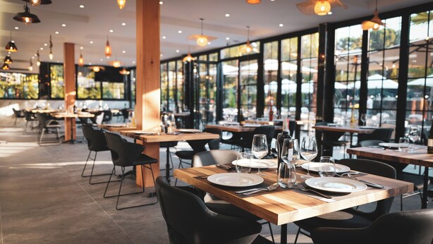 A restaurant with wooden tables and black chairs, large windows, and hanging pendant lights. - Hotel Ruko Boutique (Golem)