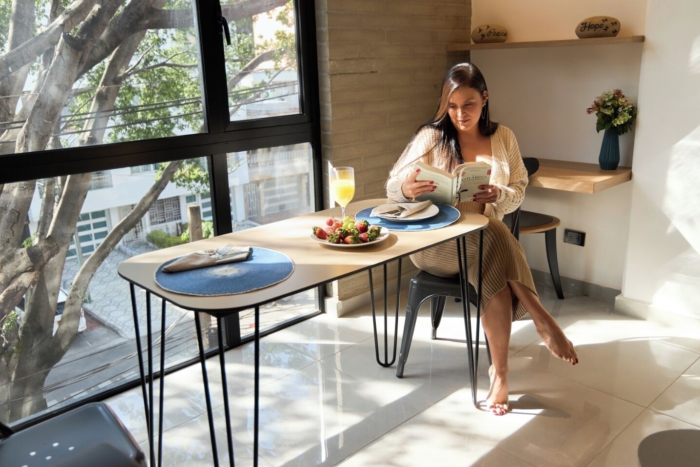 A woman reading a book at a table with a glass of orange juice, a plate of fruit, and a vase of flowers.