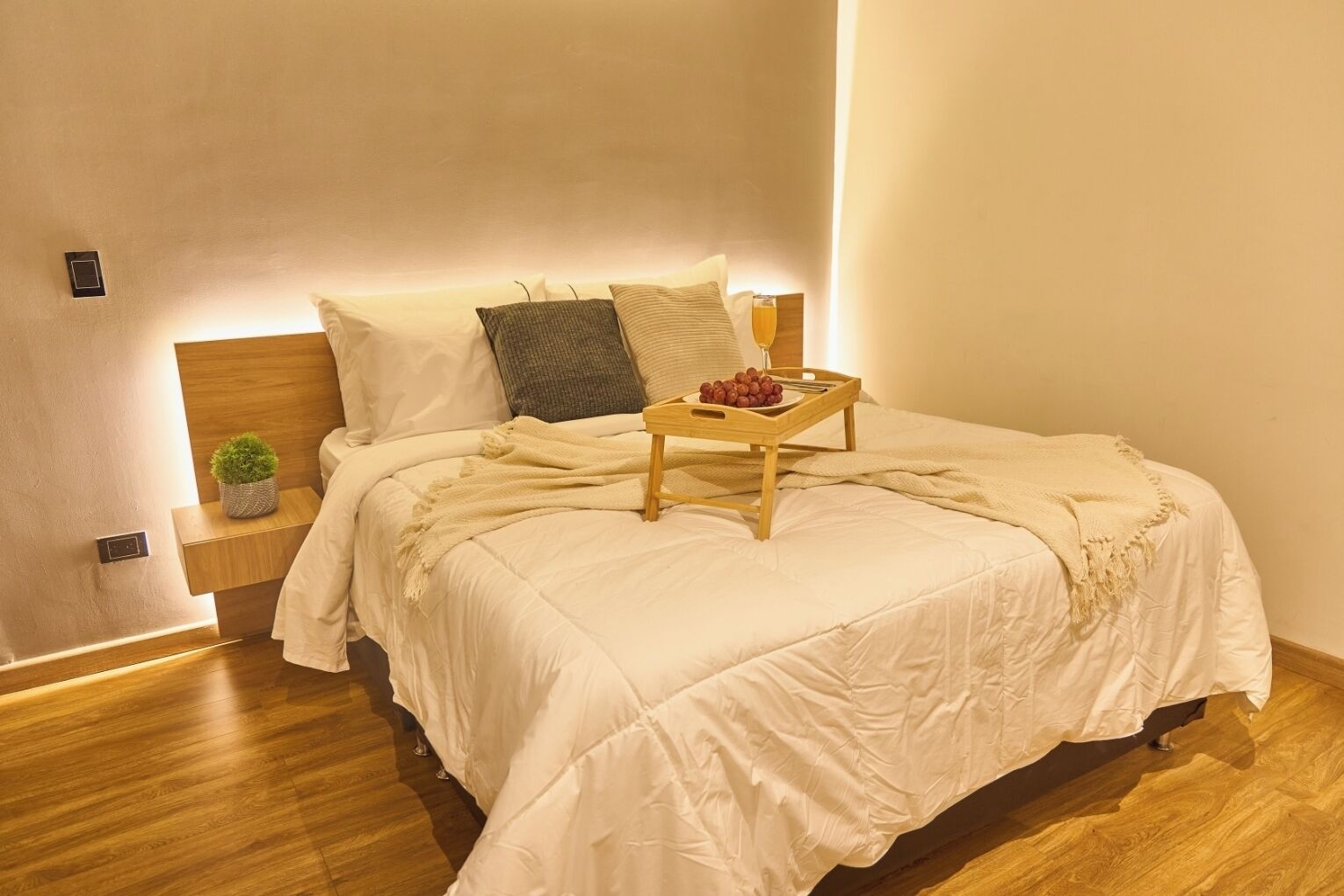 A neatly made bed with a wooden headboard, a bedside table with a lamp, and a tray with grapes and a glass.