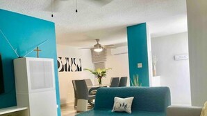 Un salon moderne avec un canapé turquoise, un meuble de rangement blanc pour les appareils électroniques et un coin repas à l’arrière-plan.