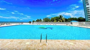 A swimming pool with clear blue water, surrounded by lounge chairs and a view of the ocean and palm trees.