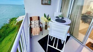 A balcony with a view of the sea, a chair, a small table with a plant, and a framed picture on the wall.