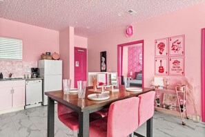 Quarto com decoração em tons de rosa, mesa de jantar posta, área de cozinha e um espelho.