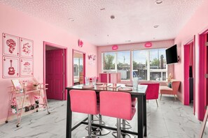 Quarto com decoração em tons de rosa, uma mesa de jantar, cadeiras, televisão e uma janela com persianas.