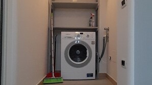 Et vaskerum med vaskemaskine, kost og en reol med genstande.
