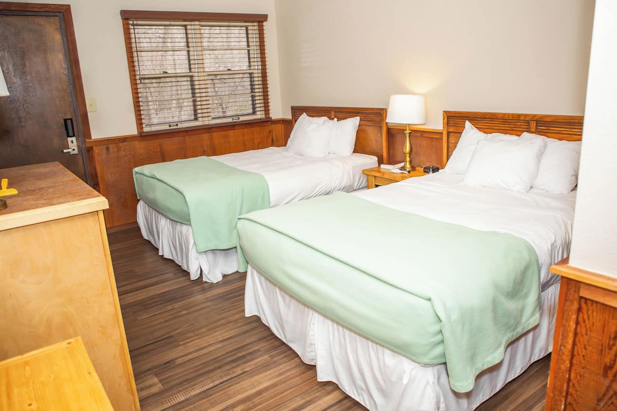 Ein Hotelzimmer mit zwei Betten, Holzkopfteilen, einem Fenster mit Jalousien, einem Nachttisch mit Lampe und einem Holztisch.
