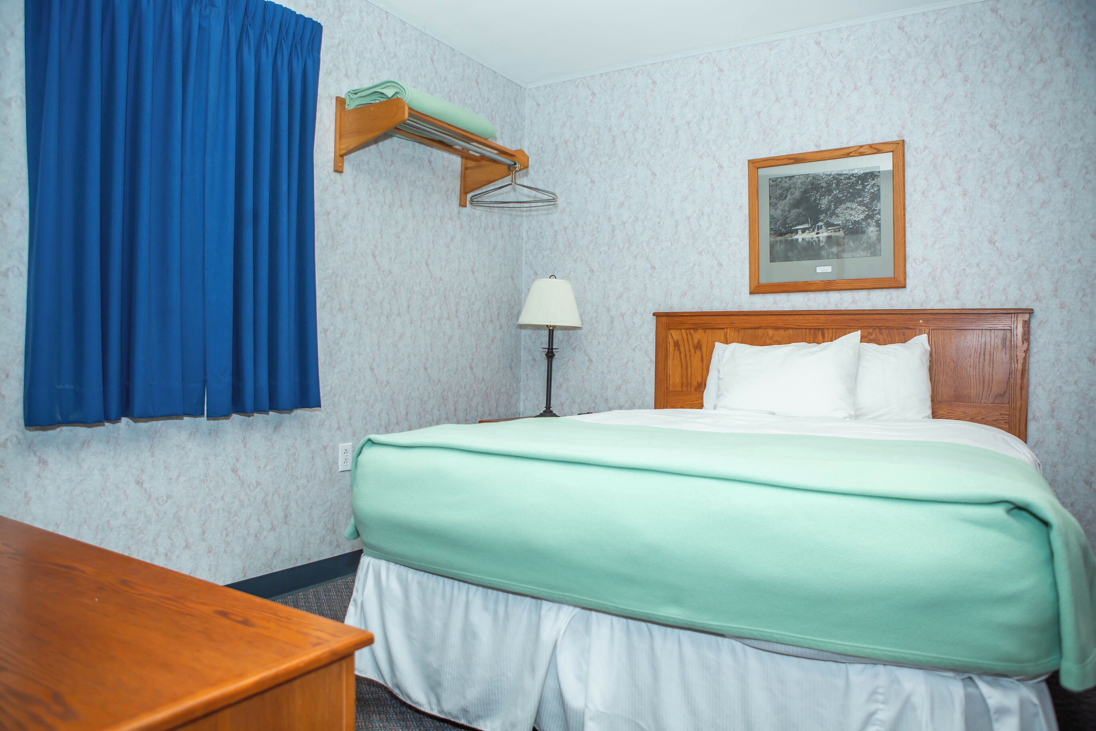 Habitación de hotel con cama, escritorio, lámpara, un cuadro en la pared y una cortina azul.