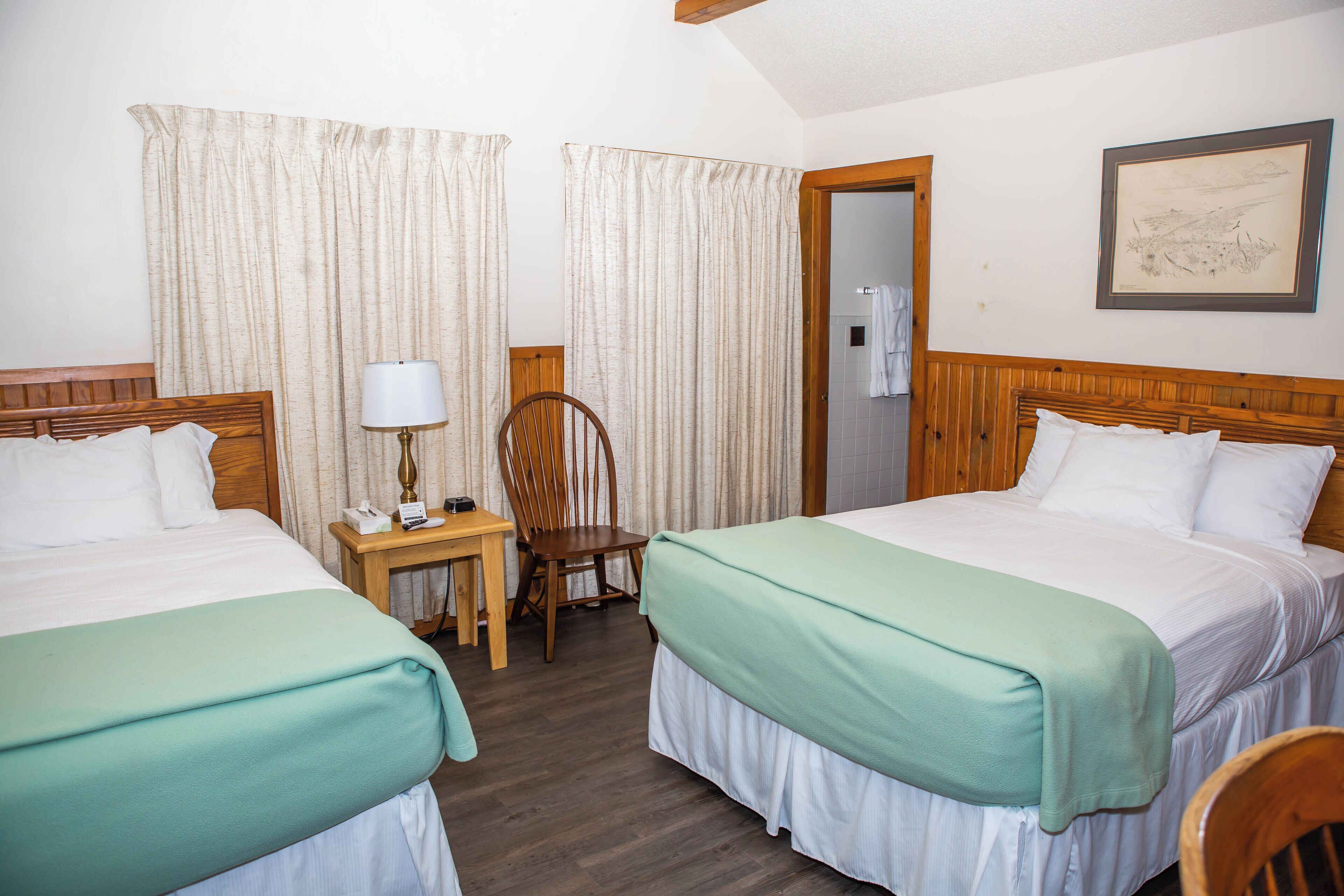 Une chambre d’hôtel avec deux lits, une table de chevet en bois avec une lampe, une chaise et une porte donnant sur une salle de bain.
