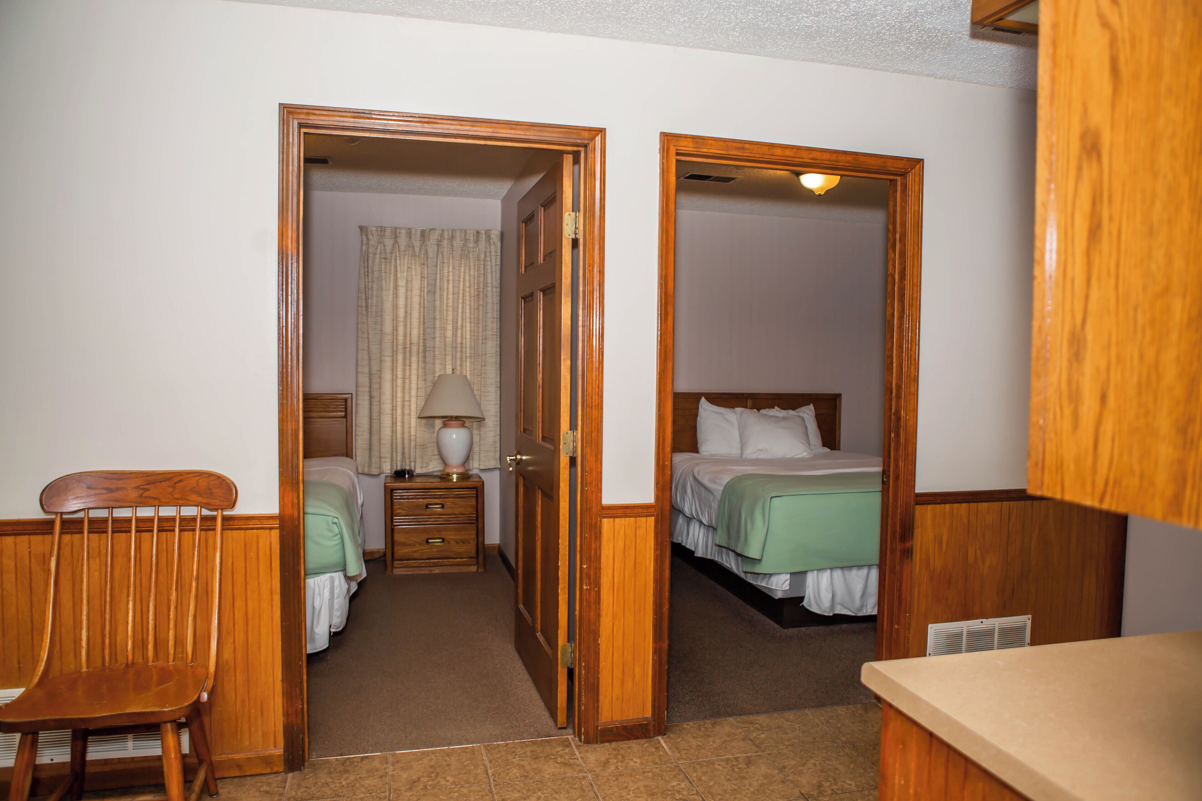 A hotel room with two beds, a wooden chair, a nightstand with a lamp, and a bathroom visible through an open door.