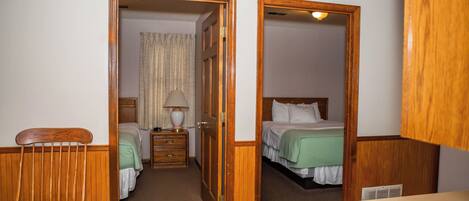 Habitación de hotel con dos camas, una silla de madera, una mesita de noche con lámpara y un baño visible a través de una puerta abierta.