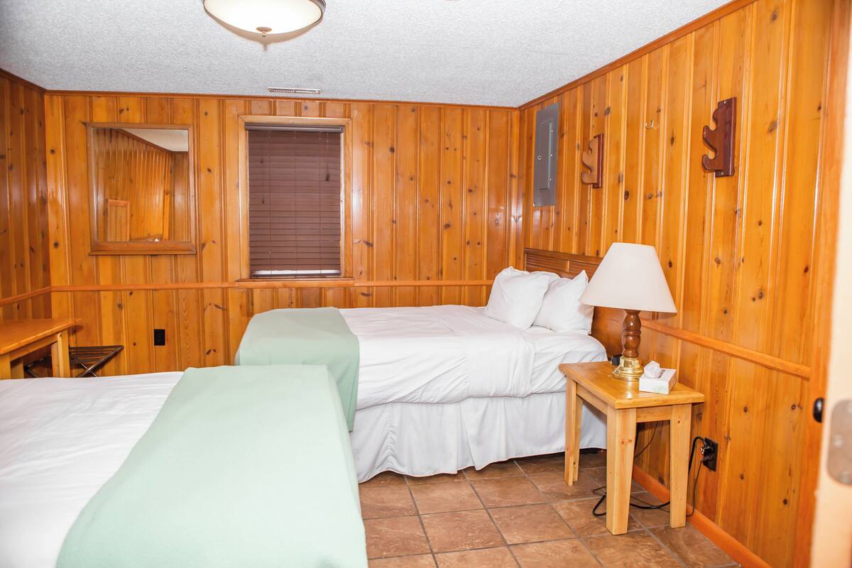 Ein Zimmer mit Holzwänden, zwei Betten, einem Nachttisch mit Lampe und einem Fenster mit Jalousien.