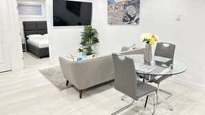 Een moderne woonkamer met een glazen eettafel, een grijze bank en een aan de muur bevestigde televisie.