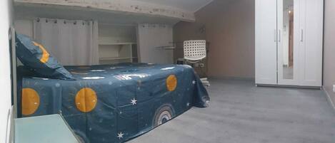 Une chambre à coucher avec un lit recouvert d’une couverture bleue ornée de cercles jaunes, une armoire blanche et une petite table.