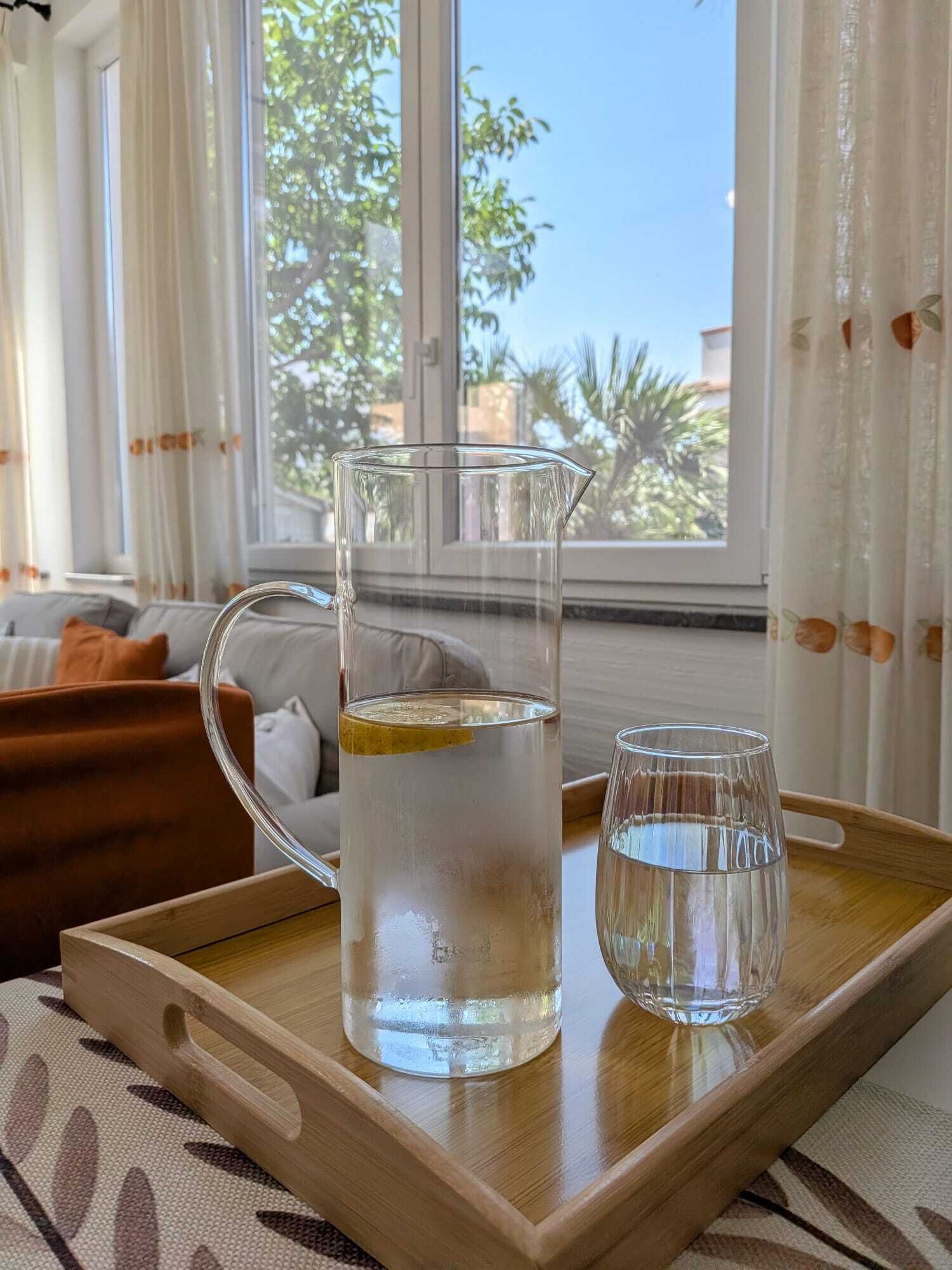 Une carafe en verre transparent, posée sur un plateau en bois avec un verre à côté, se trouve sur une table dans une pièce dotée d’une fenêtre et de rideaux.