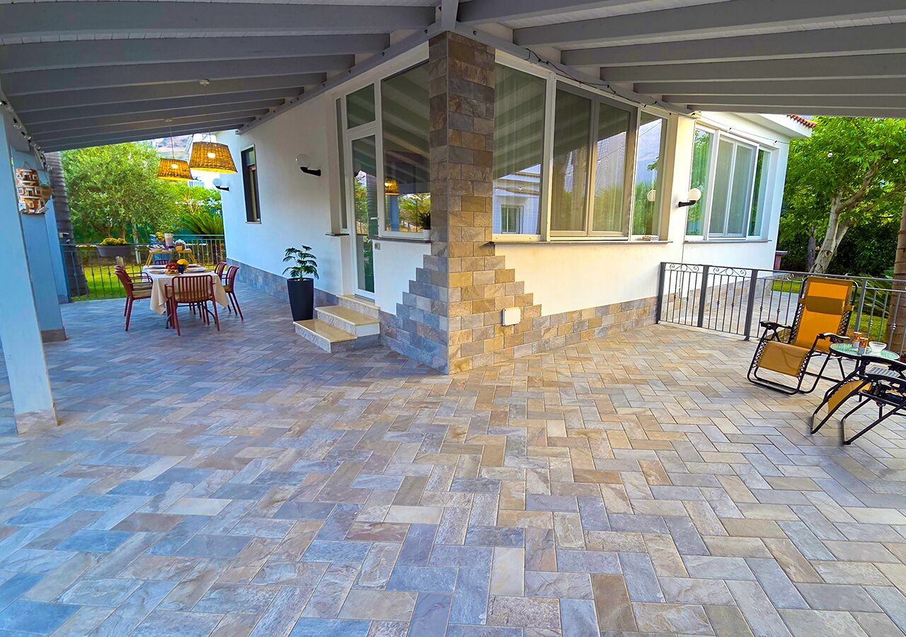 Eine überdachte Terrasse mit Steinboden, ein Essbereich mit Stühlen und Tisch sowie eine Treppe zu einer oberen Ebene.