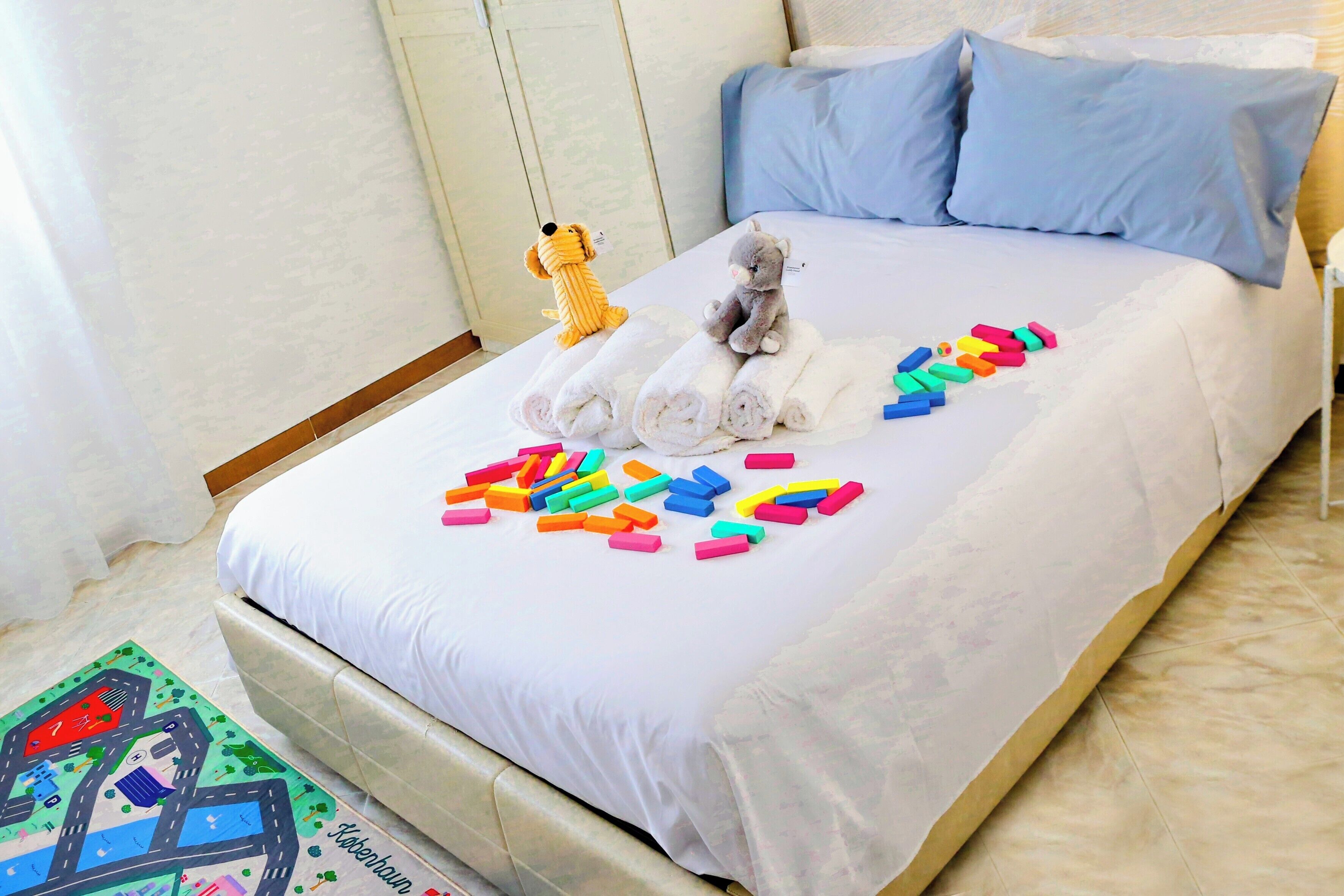 Un lit bien fait, avec un doudou et des serviettes enroulées posés dessus, un tapis coloré orné d’un motif de carte et un petit chien jouet.
