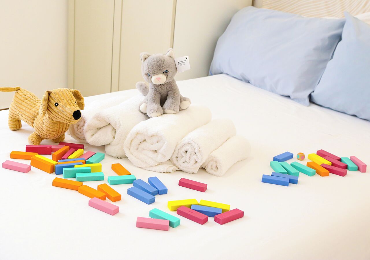 Un lit avec des serviettes blanches roulées, un ours en peluche gris et des blocs de construction colorés éparpillés.