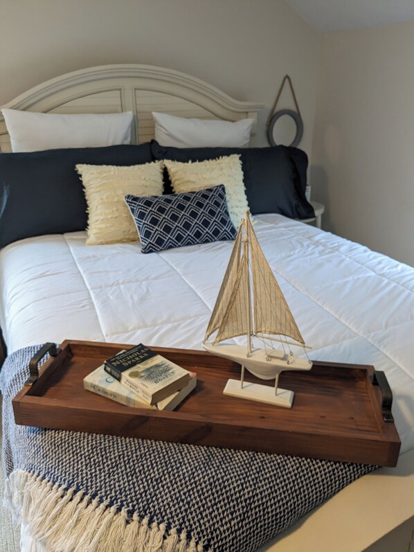 Una cama bien hecha con un modelo de velero, una bandeja con libros y una colcha de pata de gallo, sobre una cama con cabecera blanca y almohadas oscuras.