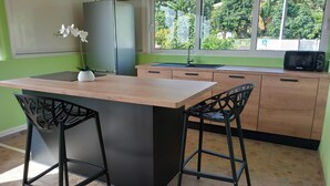 A kitchen with a central island, two bar stools, a refrigerator, a microwave, and a window with a view of greenery. - Atypical cottage ideally located for visiting the whole of Martinique.  (Rivière-Salée)