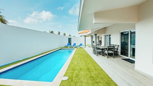 A modern outdoor pool area with a clear blue pool, a grassy lawn, and a patio with a table and chairs. - Your second home in Aruba (Oranjestad)