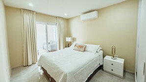 A hotel room with a large bed, a bedside table with a lamp, a nightstand, and an air conditioning unit on the wall. - Your second home in Aruba (Oranjestad)