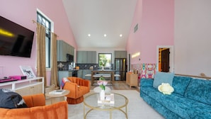 Un salon moderne avec un canapé bleu, des fauteuils orange et une table basse. Un téléviseur à écran plat est fixé au mur, et on aperçoit une cuisine au fond.