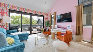 Un salon moderne doté d’une grande porte-fenêtre coulissante donnant sur un espace extérieur, d’un téléviseur à écran plat fixé au mur et de différents types de sièges, notamment un canapé bleu et un fauteuil orange.