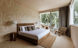 A bedroom with a large bed, a small side table, a chair, and a view of the outdoors.