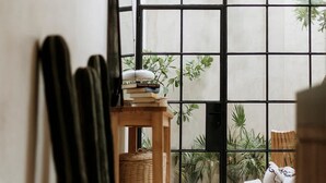Een kamer met een groot raam, een houten tafel met boeken en een plant, een bank en een cactus in een pot.