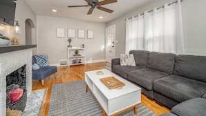 A modern living room with a fireplace, grey sofa, white coffee table, and a ceiling fan. - Game Room + Fenced Yard - King Bed - Pet Friendly (Wichita)