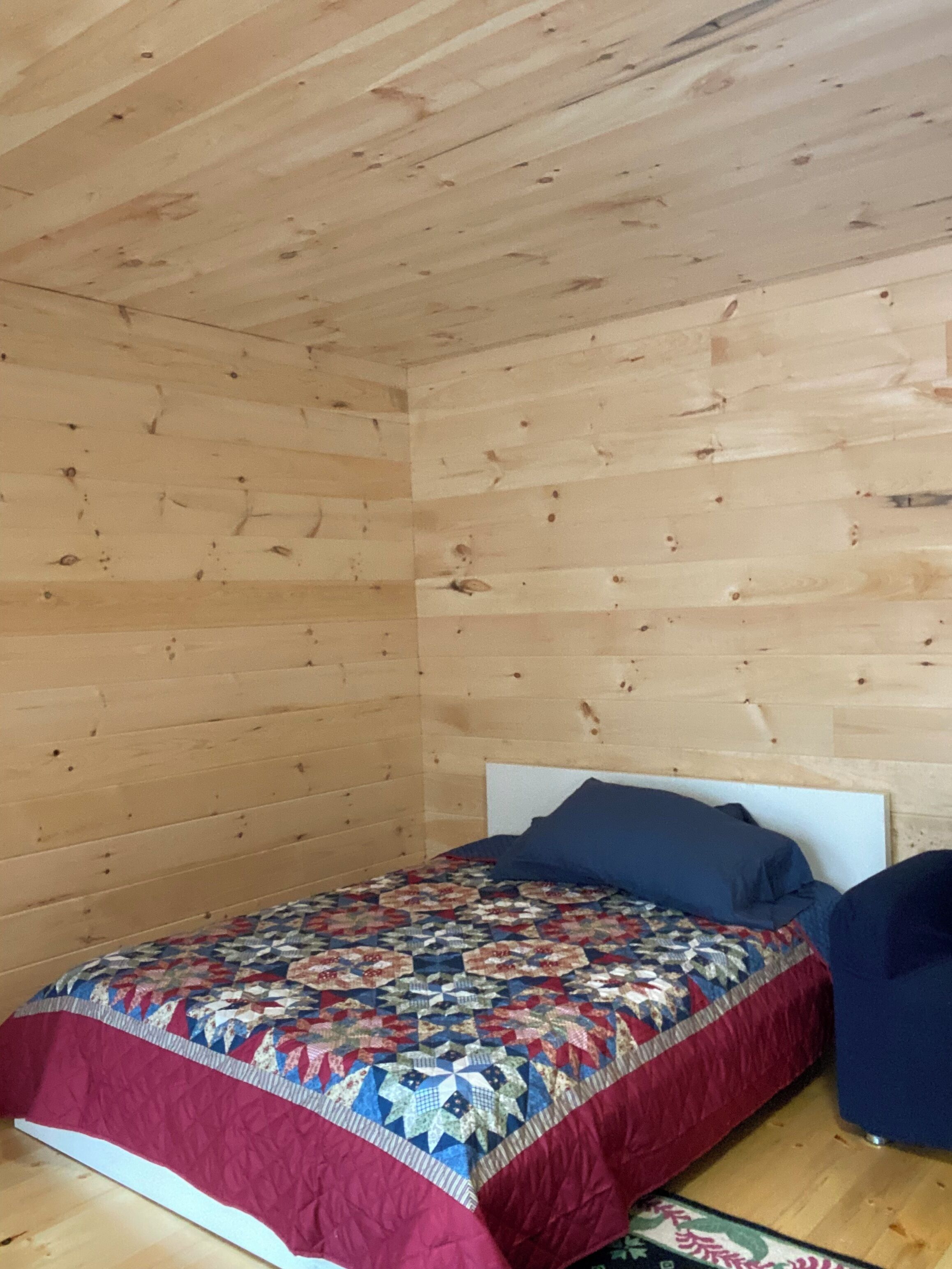 Una habitación pequeña con una cama individual, techo y paredes de madera, colcha estampada y almohada azul.