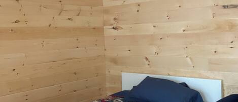 Una habitación pequeña con una cama individual, techo y paredes de madera, colcha estampada y almohada azul.
