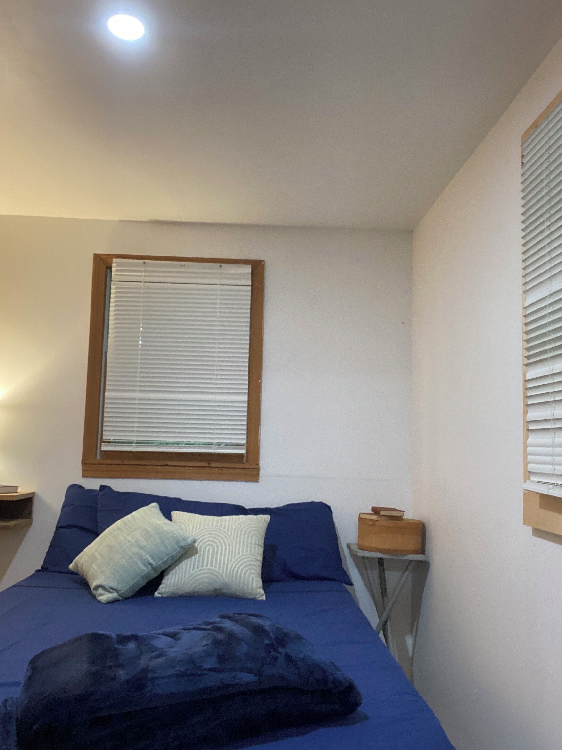 A bedroom with a bed, pillows, a bedside table, and a window with blinds.