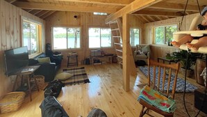 Interior de una cabaña de madera con cama en el altillo, televisor y vista a un lago a través de amplias ventanas.