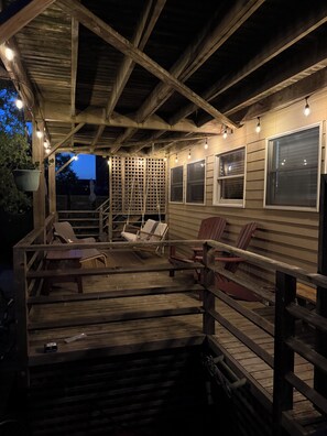 An outdoor deck with wooden railings, a seating area with chairs, and string lights. - Charming Fair Harbor 3br steps to ocean, sunset view, WiFi, A/C, outdoor shower. (Bay Shore)
