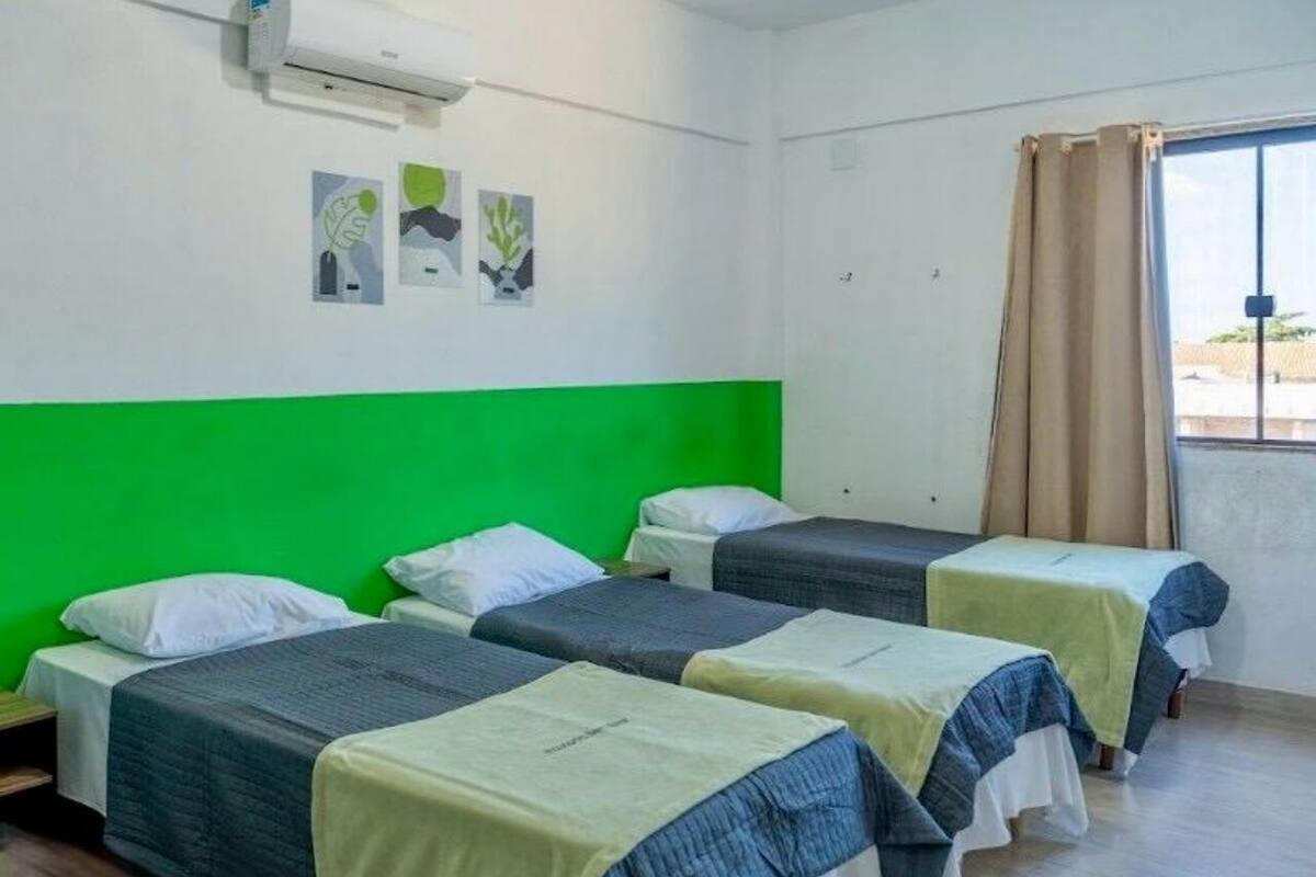 Een kamer met drie eenpersoonsbedden, elk met een groen-witte spreidek en een airconditioning op de muur, plus een raam met beige gordijnen.