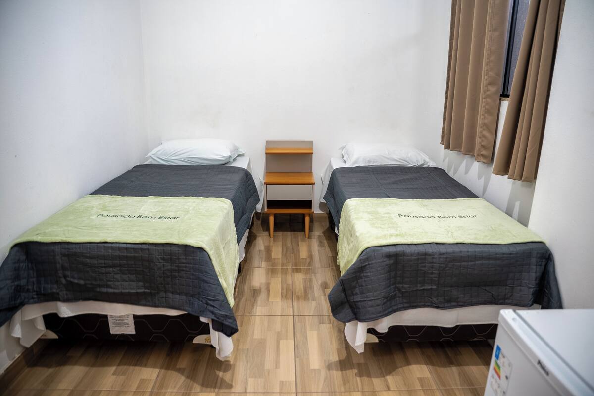Een kleine, schone kamer met twee eenpersoonsbedden, een houten nachtkastje en een raam met gordijnen.
