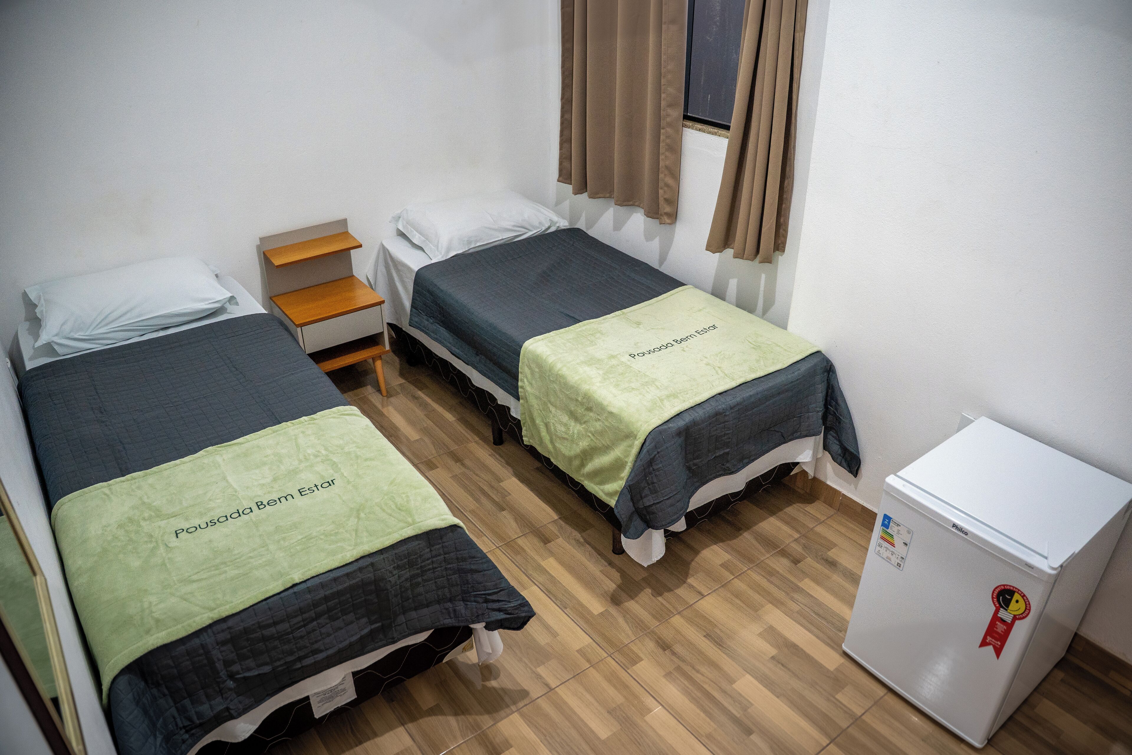 Een studentenkamer met twee eenpersoonsbedden, een klein houten nachtkastje, een witte koelkast en een raam met gordijnen.
