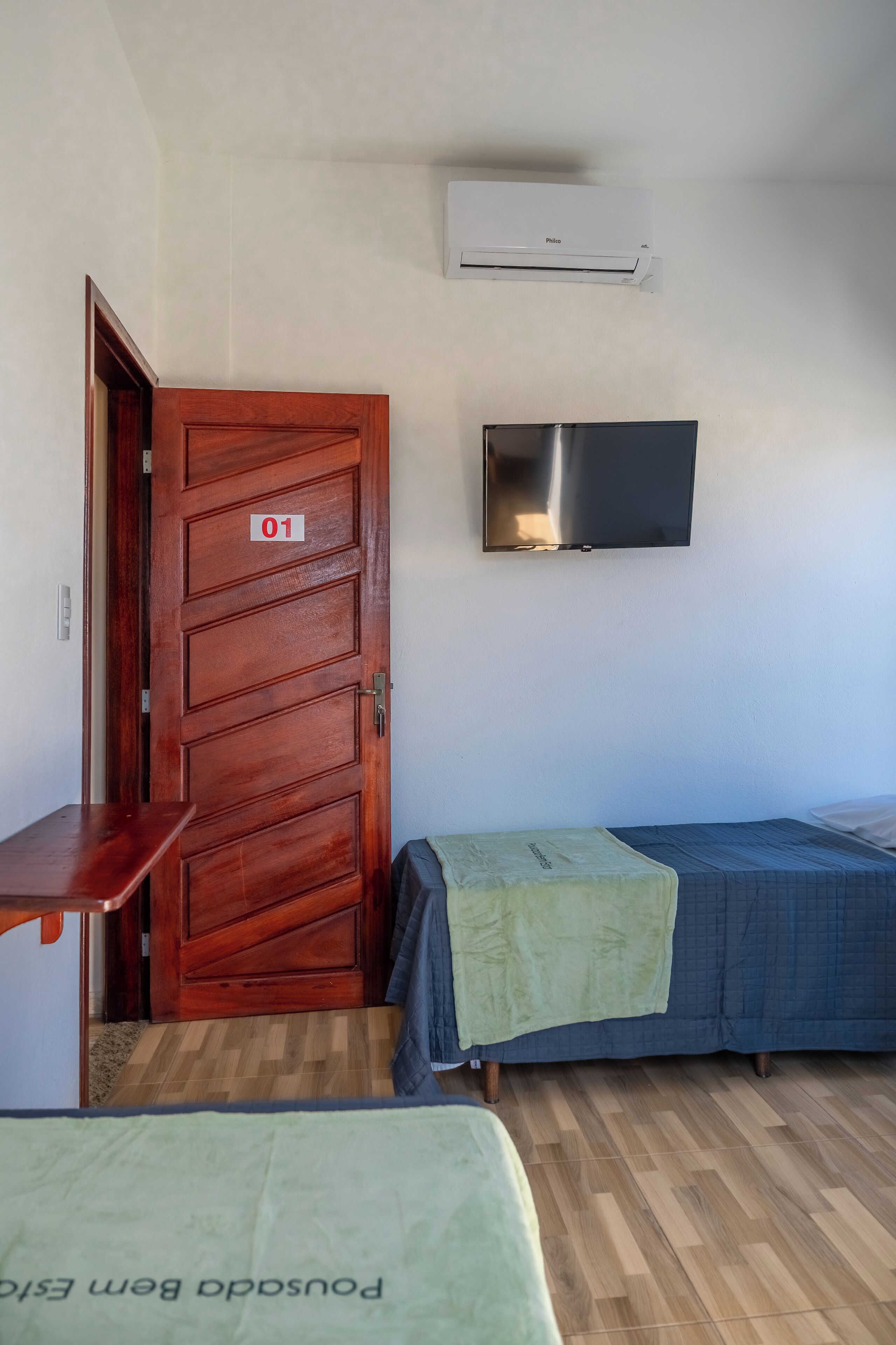 Quarto com porta de madeira identificada com o número 01, televisão de tela plana na parede e duas camas com roupa de cama verde e azul.