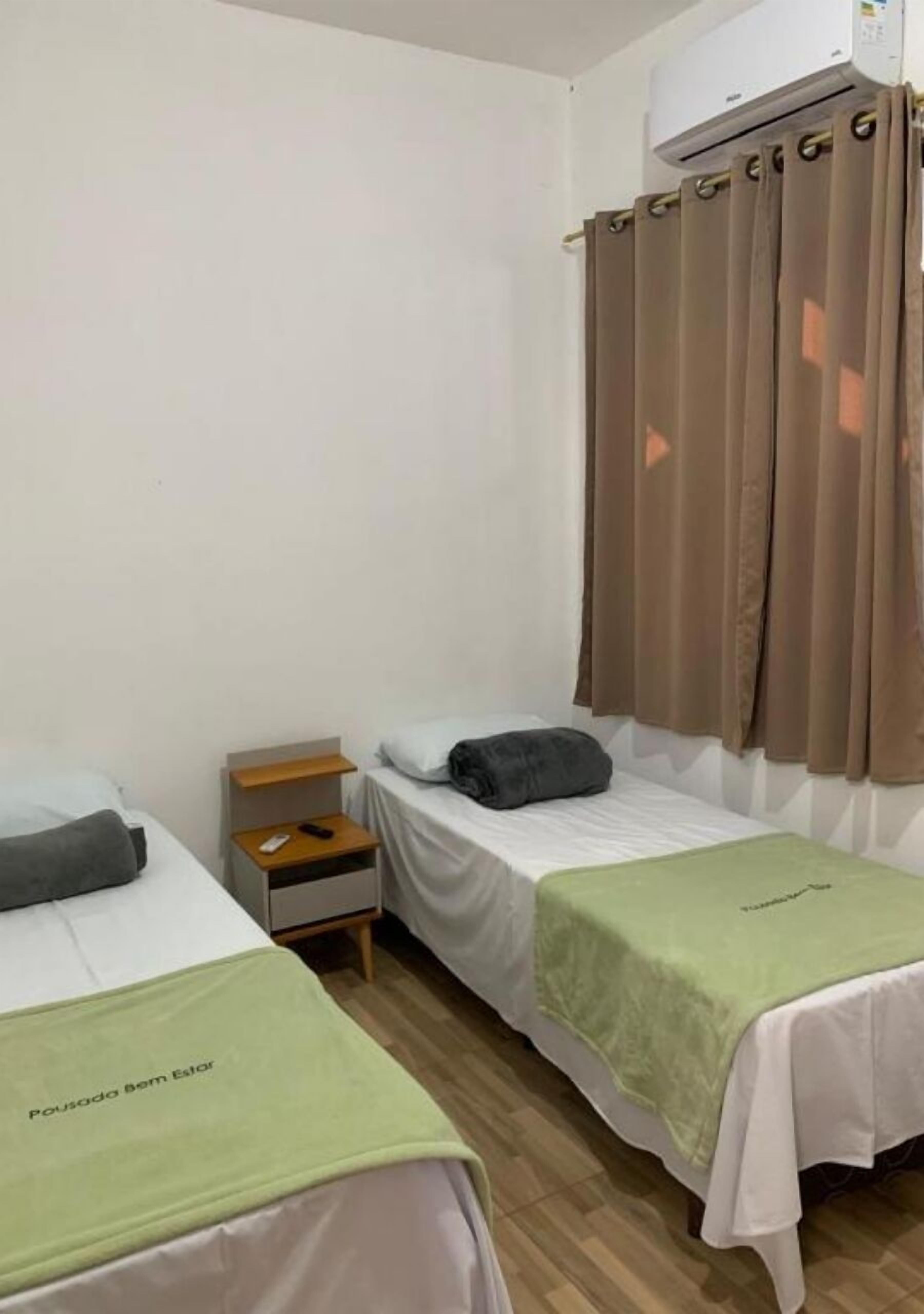 Una stanza piccola e pulita con due letti singoli, un comodino in legno, un asciugamano verde e una finestra con tende marroni.