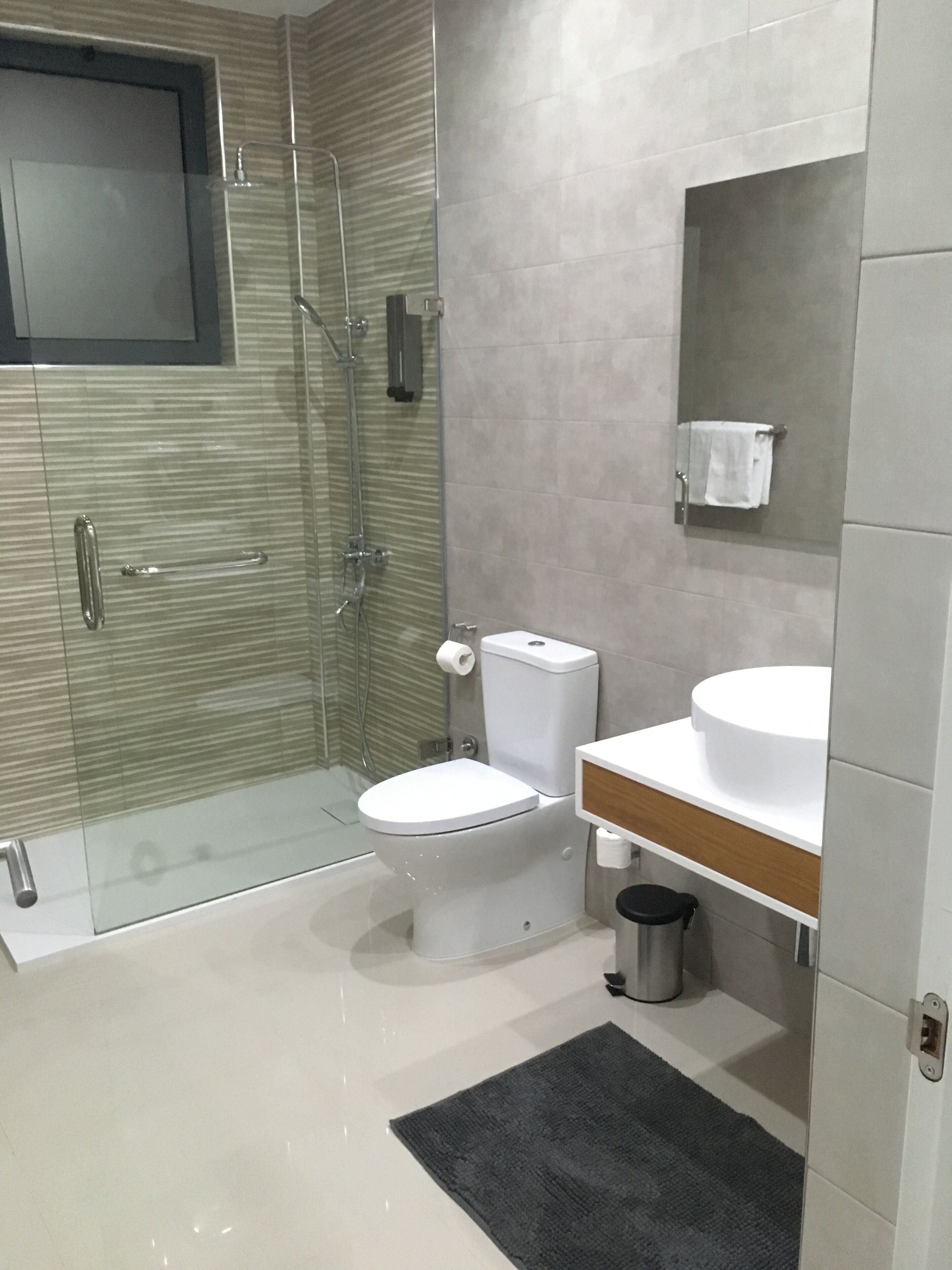 Une salle de bain moderne avec une douche à parois de verre, des toilettes blanches, un lavabo avec un plan de travail en bois et une petite poubelle.