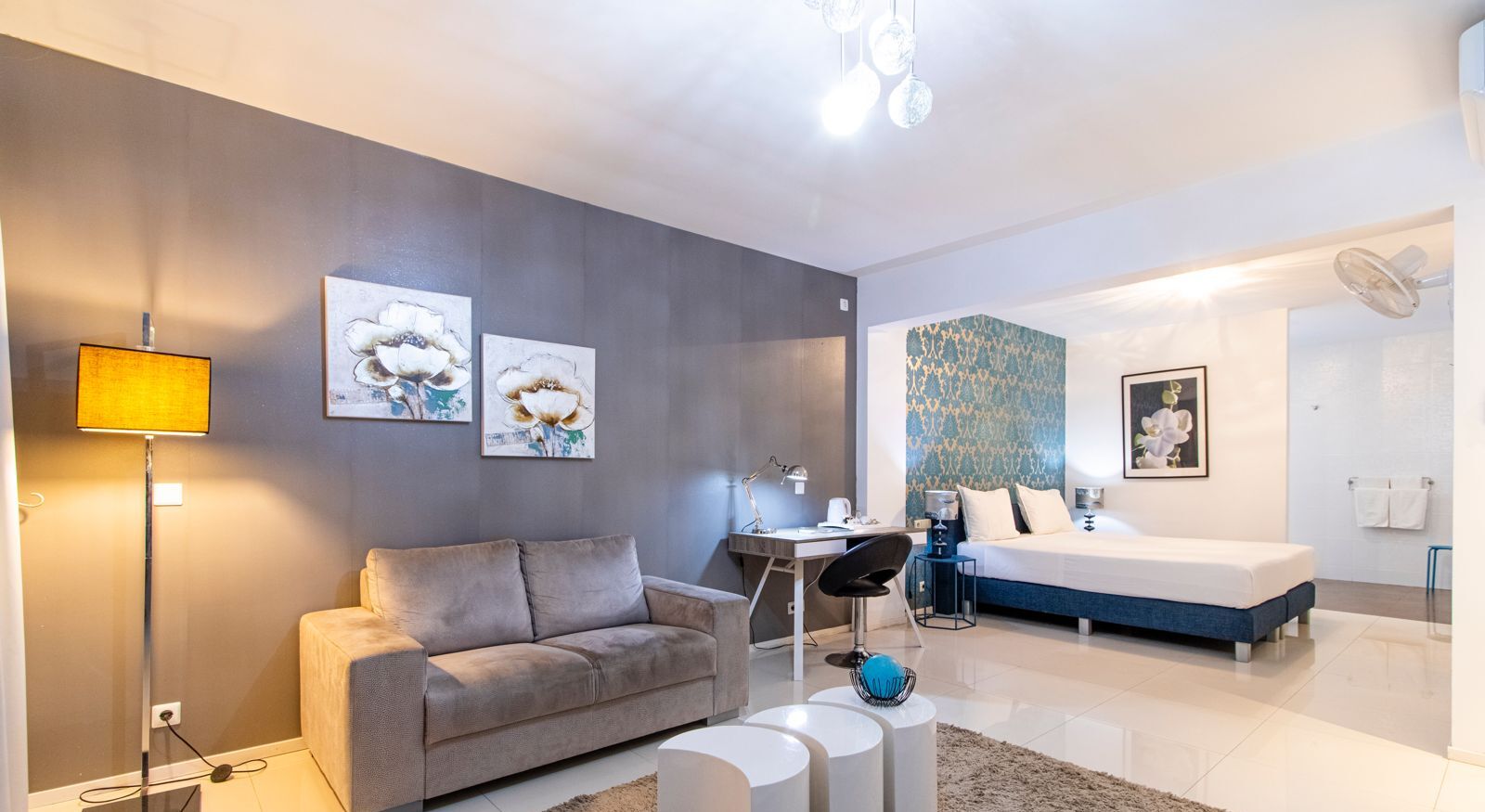 Une chambre d’hôtel moderne avec un canapé gris, un bureau, un lit avec une tête de lit bleue et des œuvres d’art accrochées aux murs.