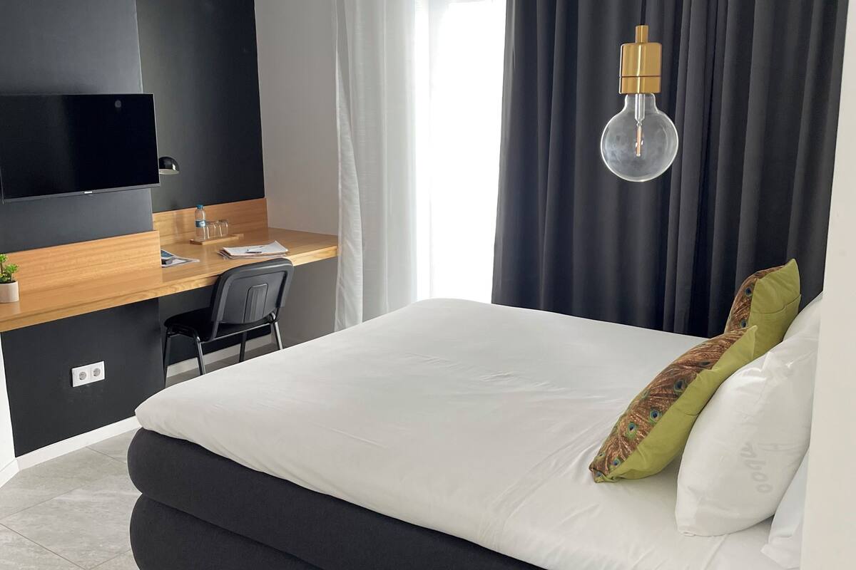 A modern hotel room with a large bed, a desk with a chair, a flat-screen TV, a telephone, and a window with curtains.