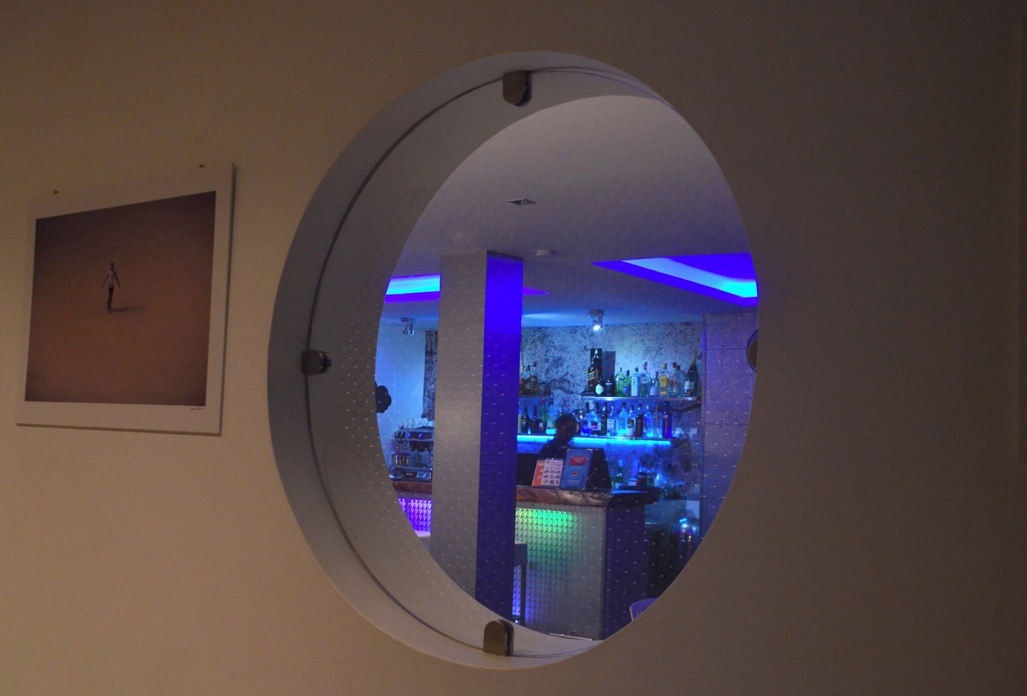 A circular window with a bar visible through it, featuring blue lighting and a variety of bottles.