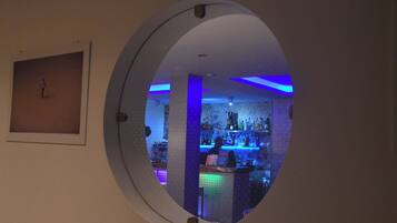 A circular window with a bar visible through it, featuring blue lighting and a variety of bottles.