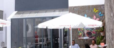 An outdoor café with tables and chairs, a glass enclosure, and a mural on the wall.