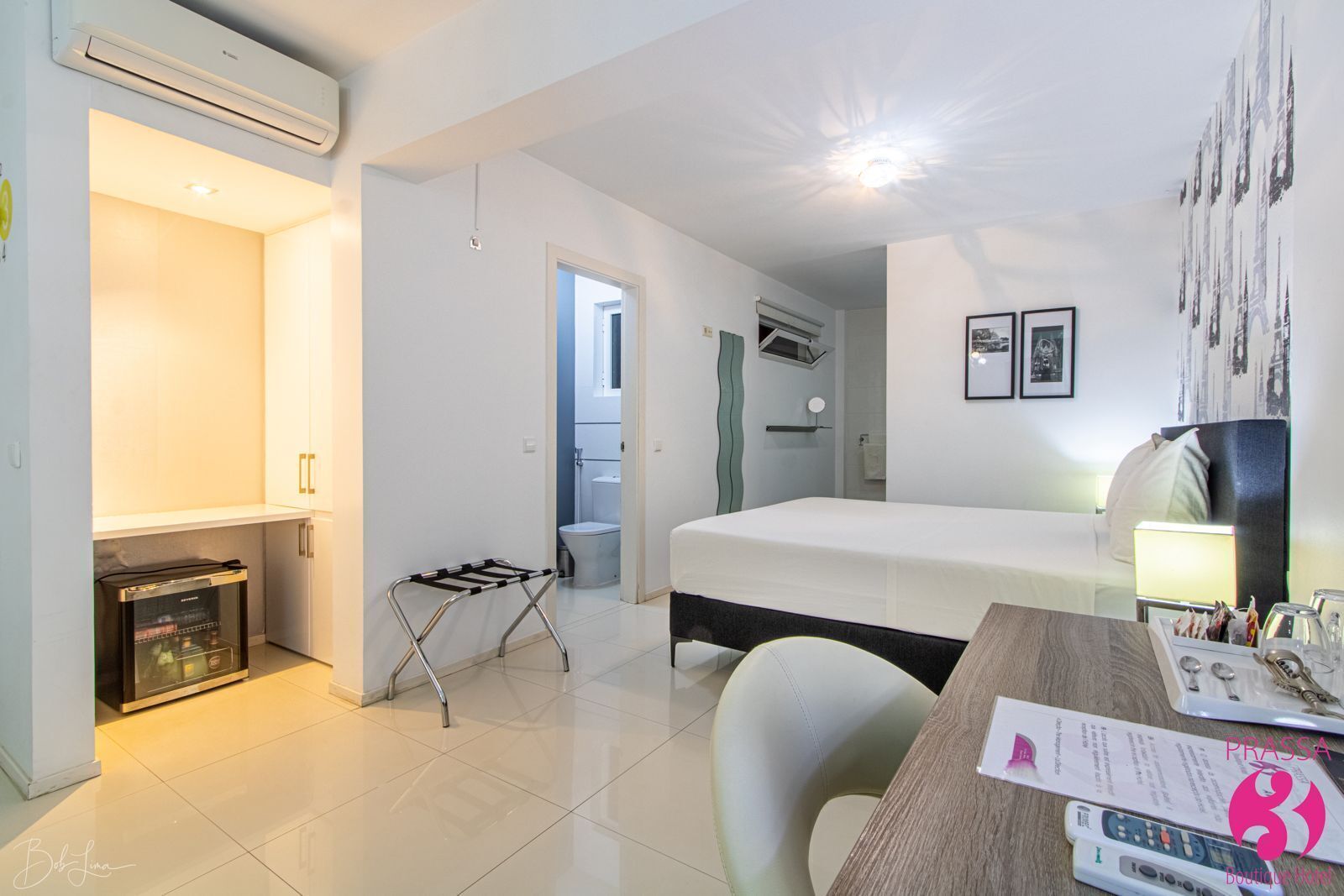 Une chambre d’hôtel moderne équipée d’un lit, d’un bureau avec une lampe, d’une petite kitchenette et d’une salle de bain visible à travers une porte ouverte.
