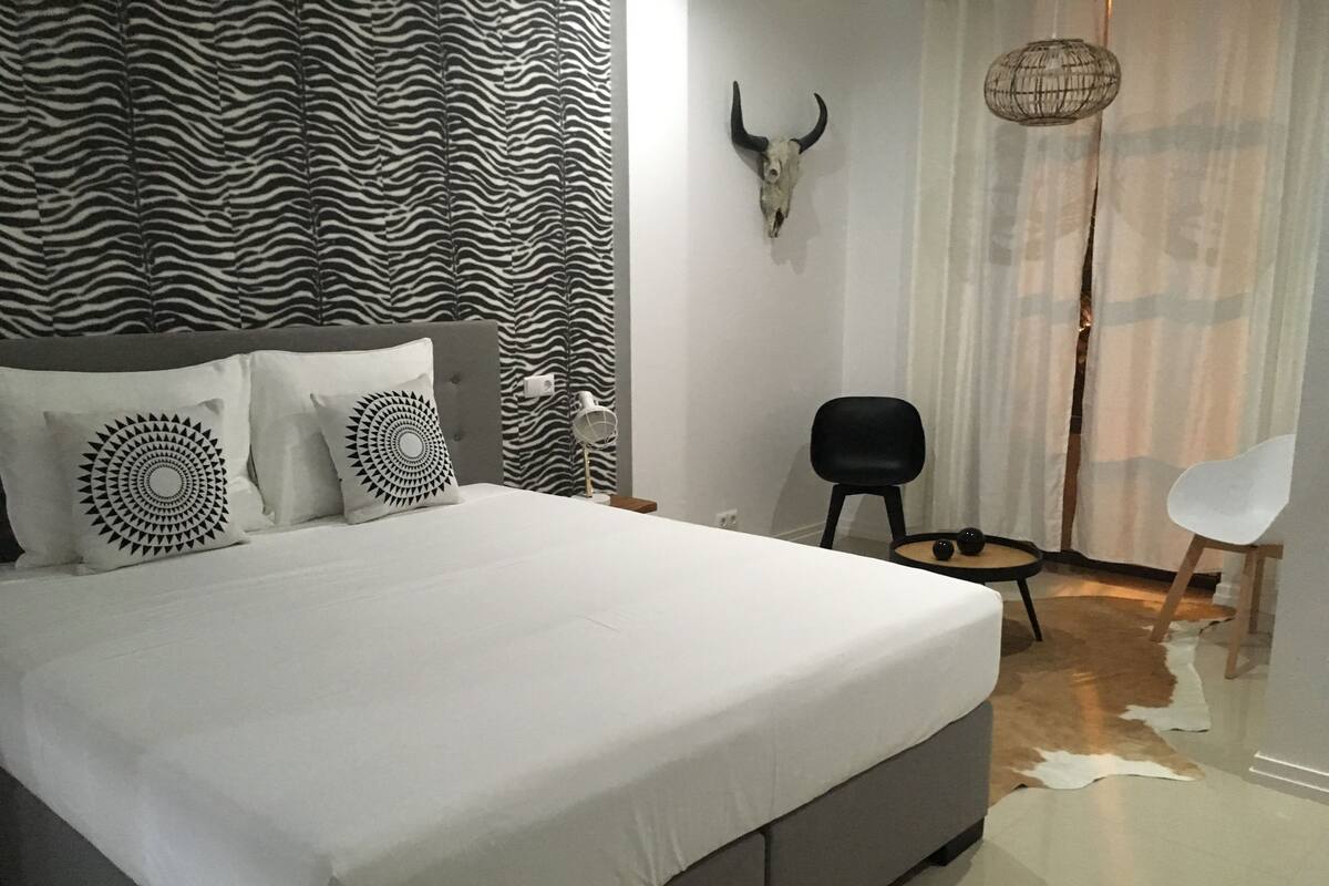 A bedroom with a zebra-patterned headboard, a bed with white bedding, a black chair, a white chair, and a wall-mounted bull skull.