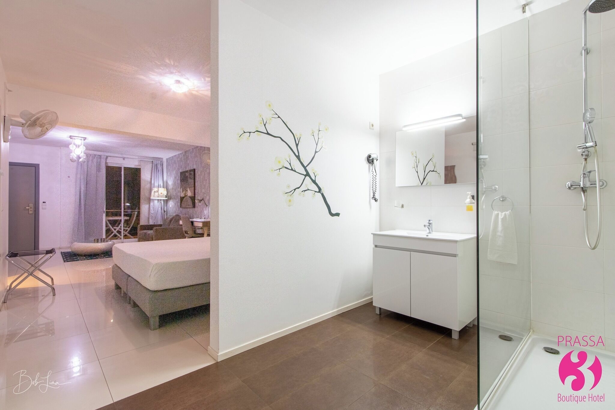 Een moderne hotelkamer met een groot bed, een zithoek met een stoel en tafel, een badkamer met douche, en een spiegel aan de muur.