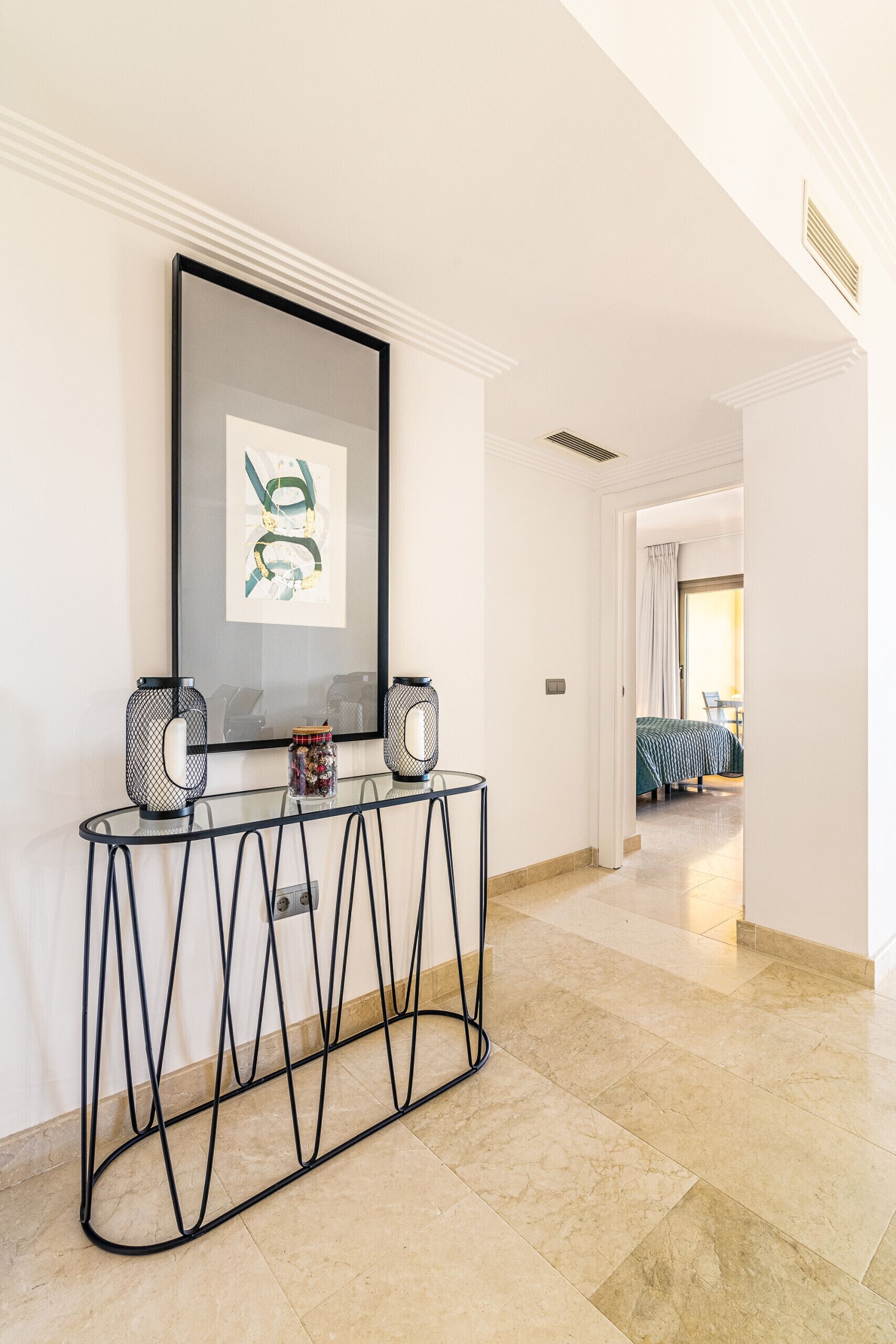 A hallway with a framed artwork, a console table with two speakers, and a view into a bedroom with a bed and curtains.